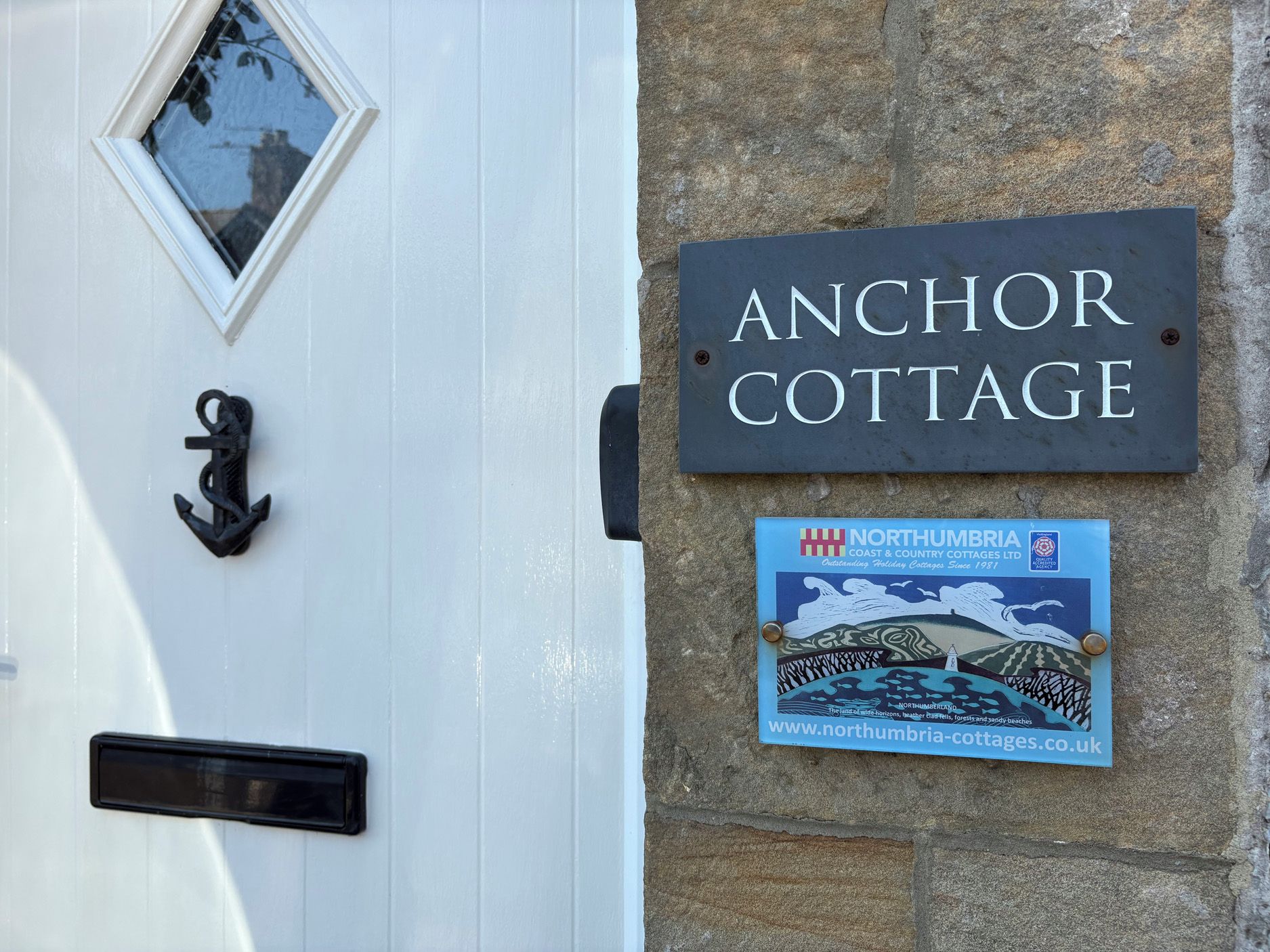 A white door with a black mailbox and a black door knocker. A sign above the door reads 'Anchor Cottage.'