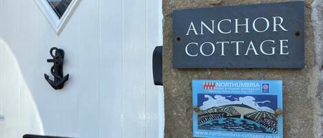 A white door with a black mailbox and a black door knocker. A sign above the door reads 'Anchor Cottage.'