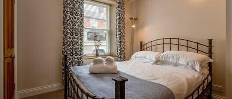 A neatly made bed with a headboard, a bedside lamp, and a window with patterned curtains.
