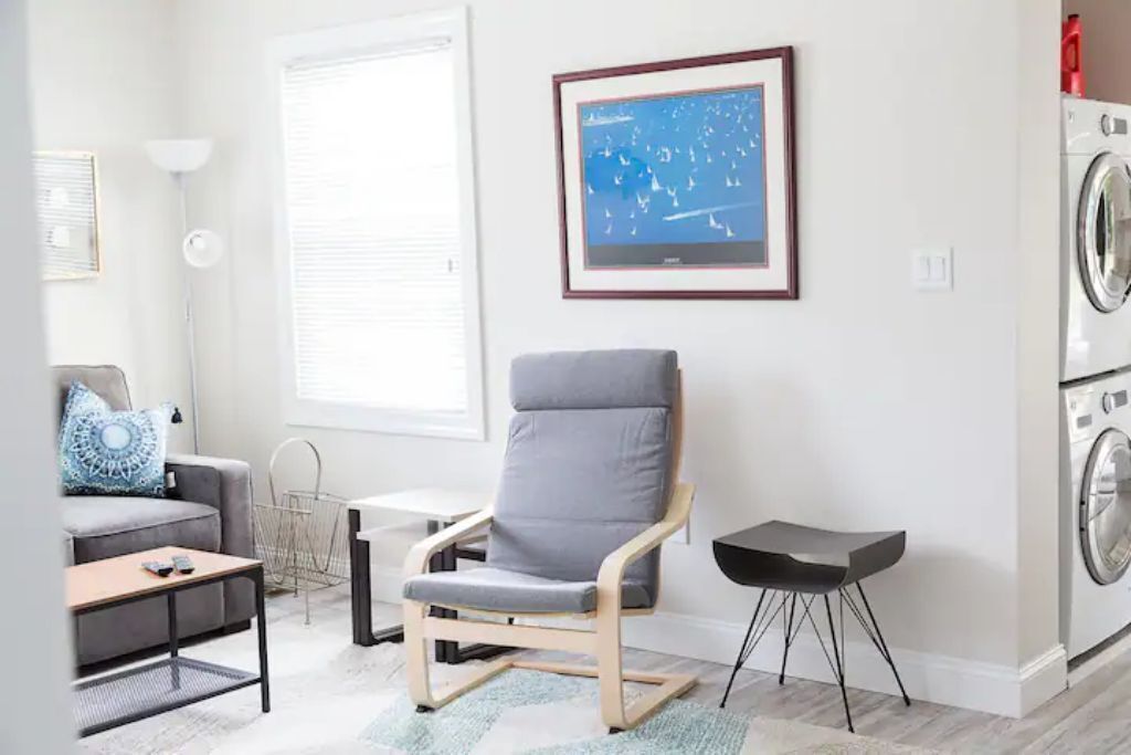 Una sala moderna con un sillón gris, un sofá gris, una mesita y un cuadro enmarcado en la pared. Se observa una lavadora al fondo.