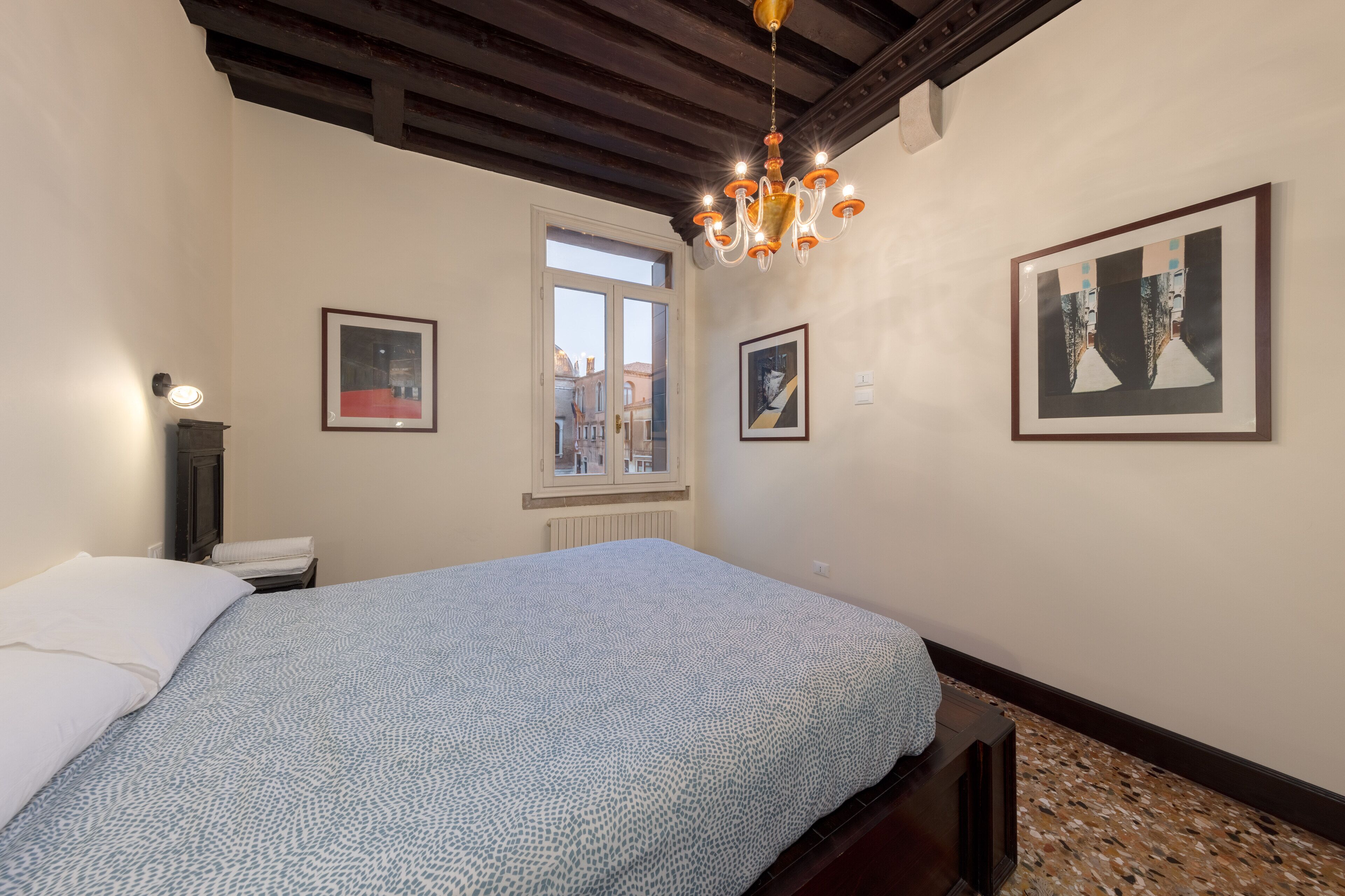 Un dormitorio con cama, ventana, candelabro y cuadros en la pared.
