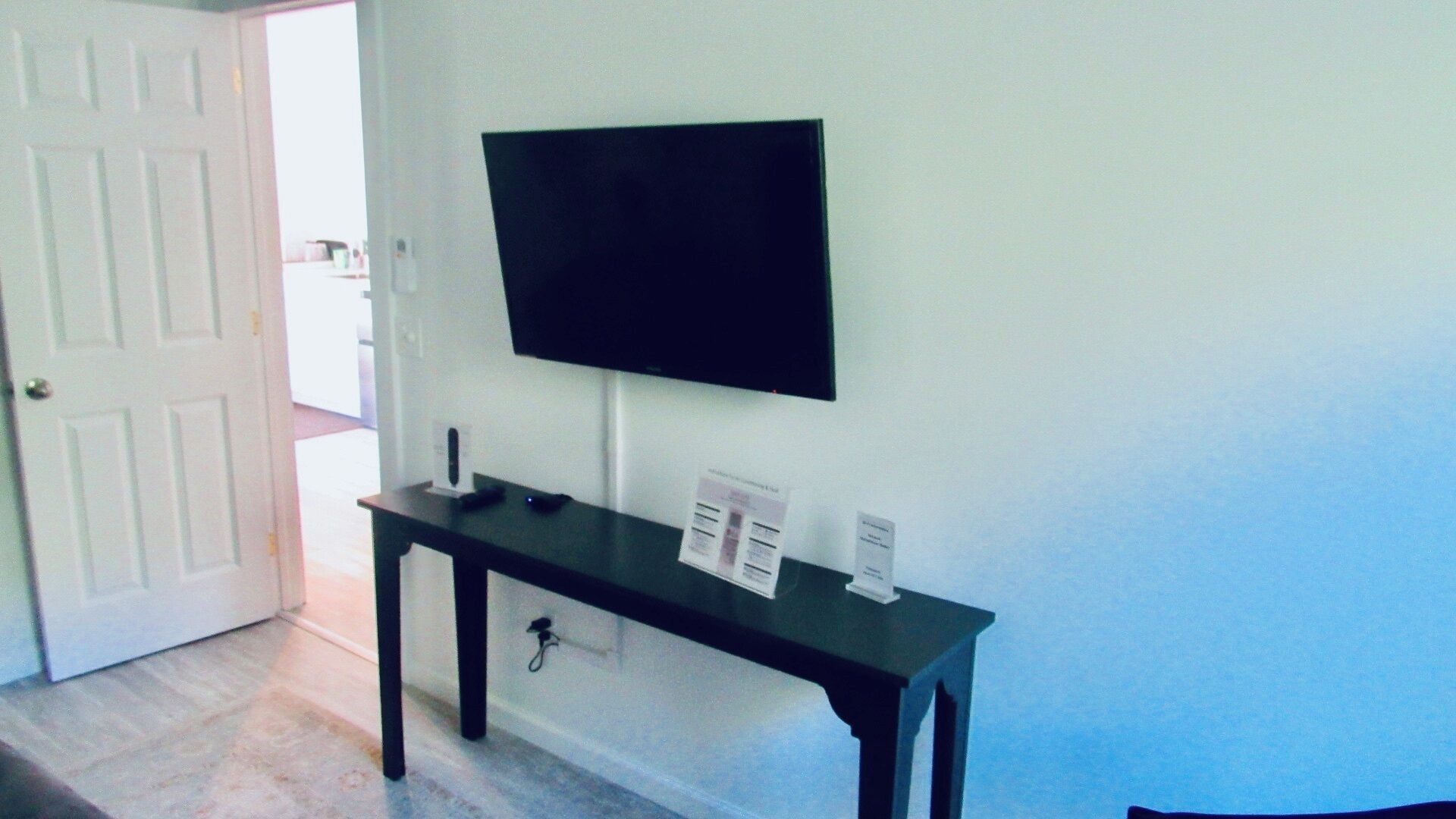 Una habitación con una mesa de madera oscura, un televisor de pantalla plana montado en la pared y una puerta que da a otra habitación.