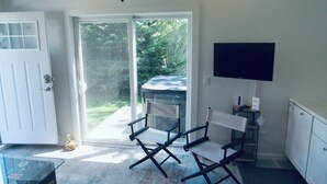 A room with a sliding glass door leading to a garden, a small table, a chair, a television mounted on the wall, and a cabinet. - Blue Bay Dream w/ Seasonal Outdoor Jacuzzi, Cabana + Office (Greenport,)