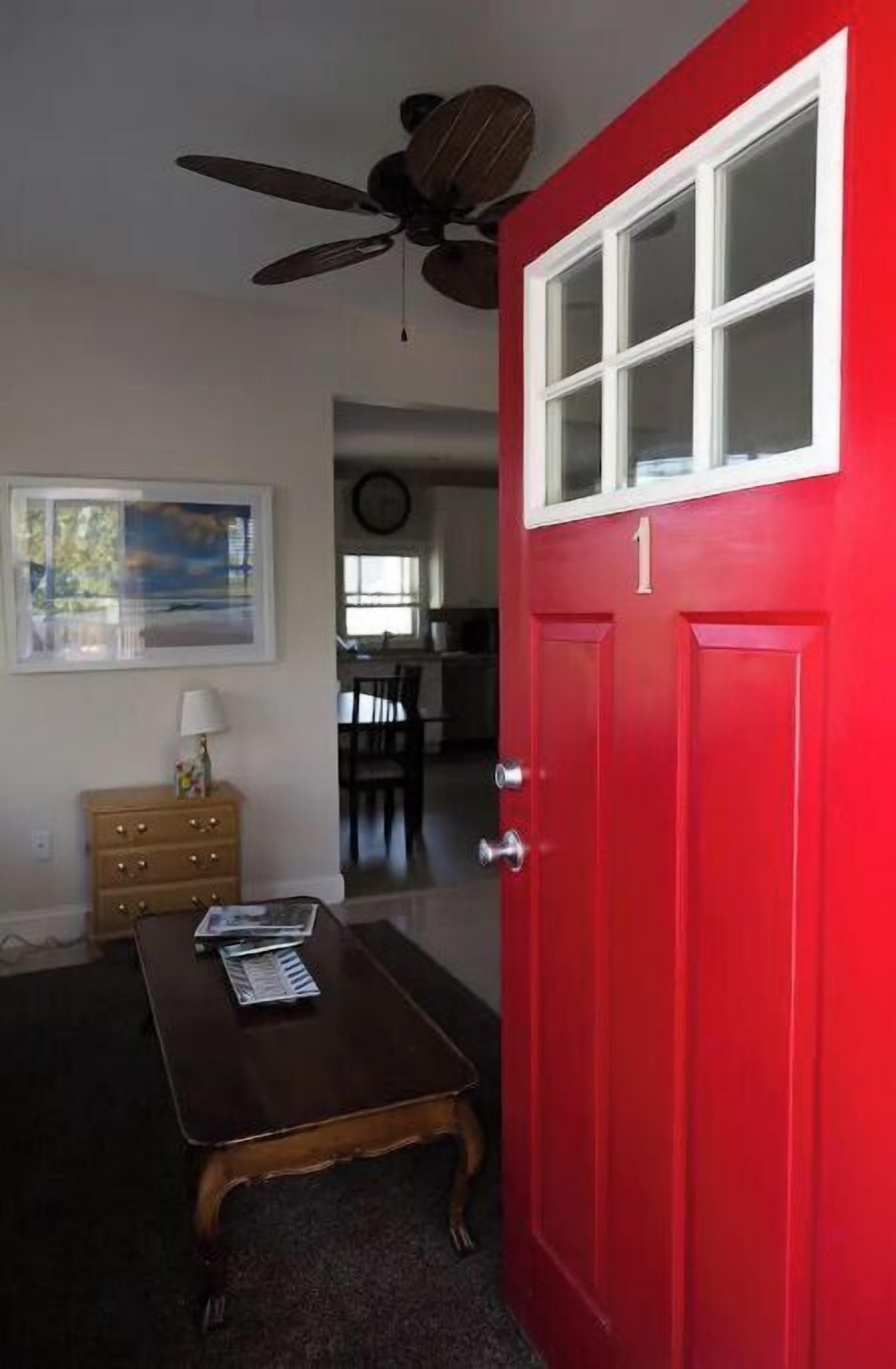 A room with a red door, a ceiling fan, a wooden table, a dresser, and a painting on the wall.