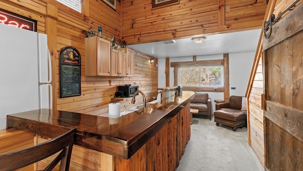 A wooden cabin interior with a kitchen area featuring a wooden bar, a refrigerator, a sink, and a seating area with two chairs and a sofa. - "The Lodge"-  APT w/Loft Bedroom, Walnut Bar, Quiet Setting, Covered Parking (Enterprise)