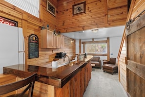 A wooden cabin interior with a kitchen area featuring a wooden bar, a refrigerator, a sink, and a seating area with two chairs and a sofa.