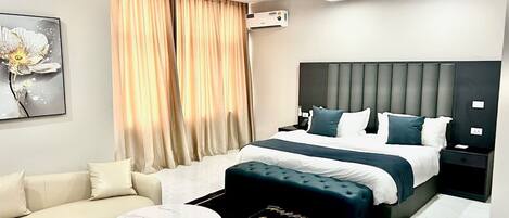 A modern hotel room with a large bed, a sofa, a round coffee table, and a decorative ottoman.