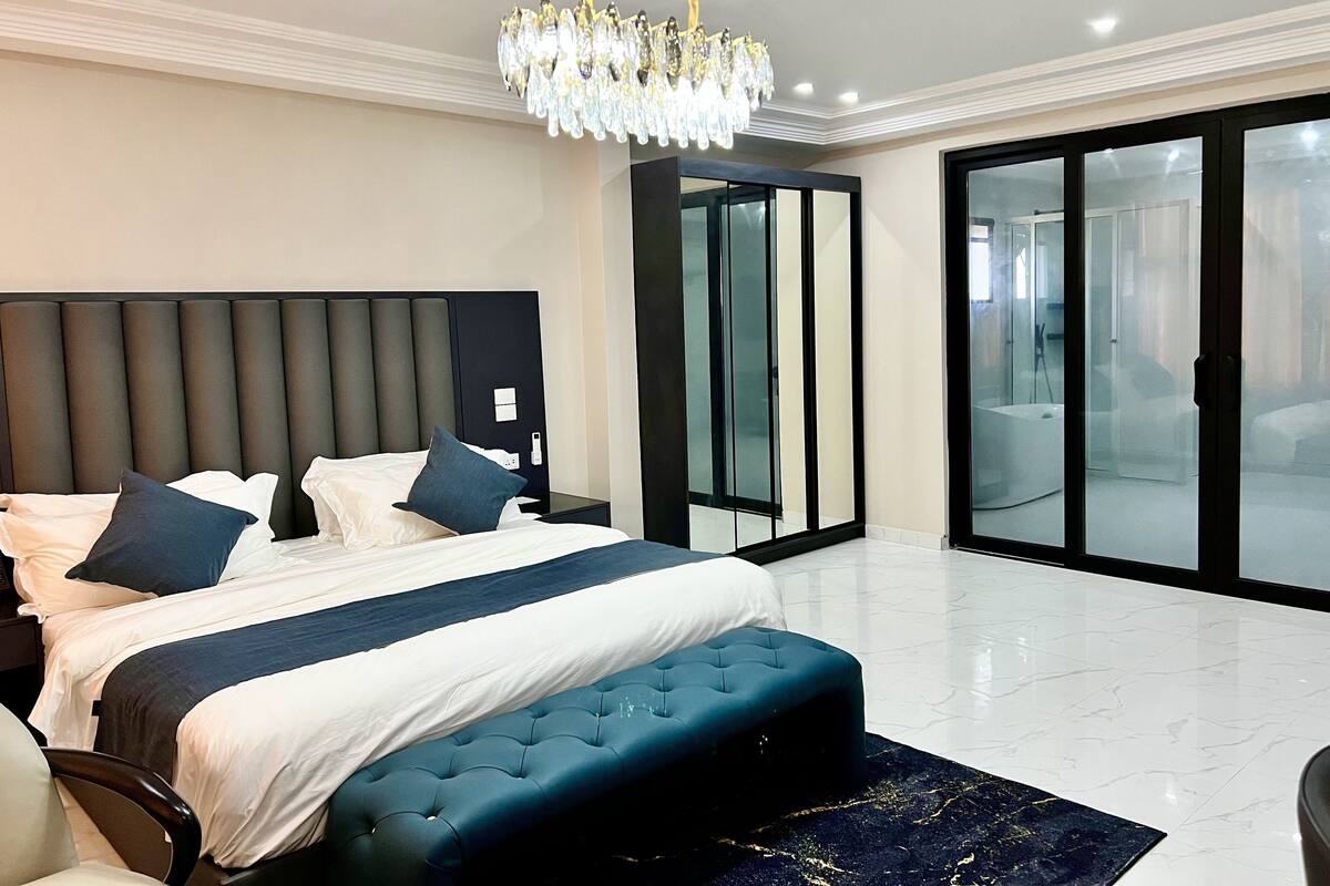 Een moderne slaapkamer met een groot bed, een gestofte bank, een kroonluchter en een glazen schuifdeur naar een badkamer.