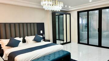 Een moderne slaapkamer met een groot bed, een gestofte bank, een kroonluchter en een glazen schuifdeur naar een badkamer.