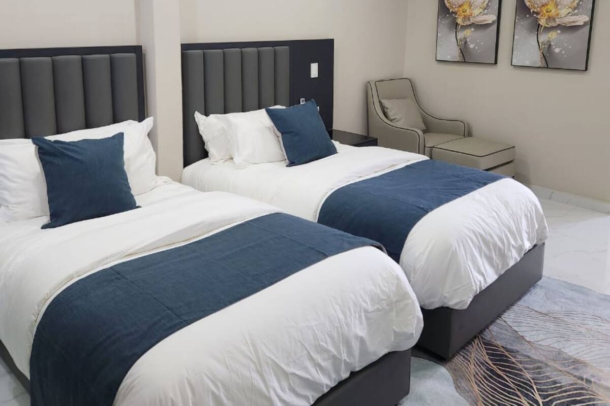Een moderne hotelkamer met twee bedden, een grijze hoofdbord, een stoel en twee bloemschilderijen aan de muur.
