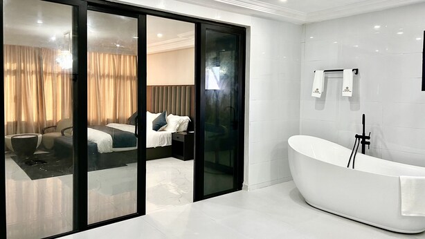 Una habitación de hotel moderna con un gran ventanal que da a un dormitorio, una bañera blanca y un espejo grande.