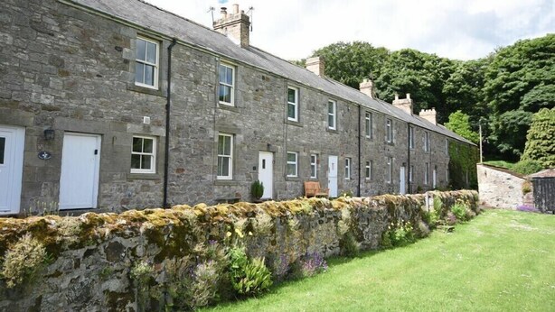 A row of stone buildings with multiple windows and doors, a stone wall in front, and a grassy area with plants.