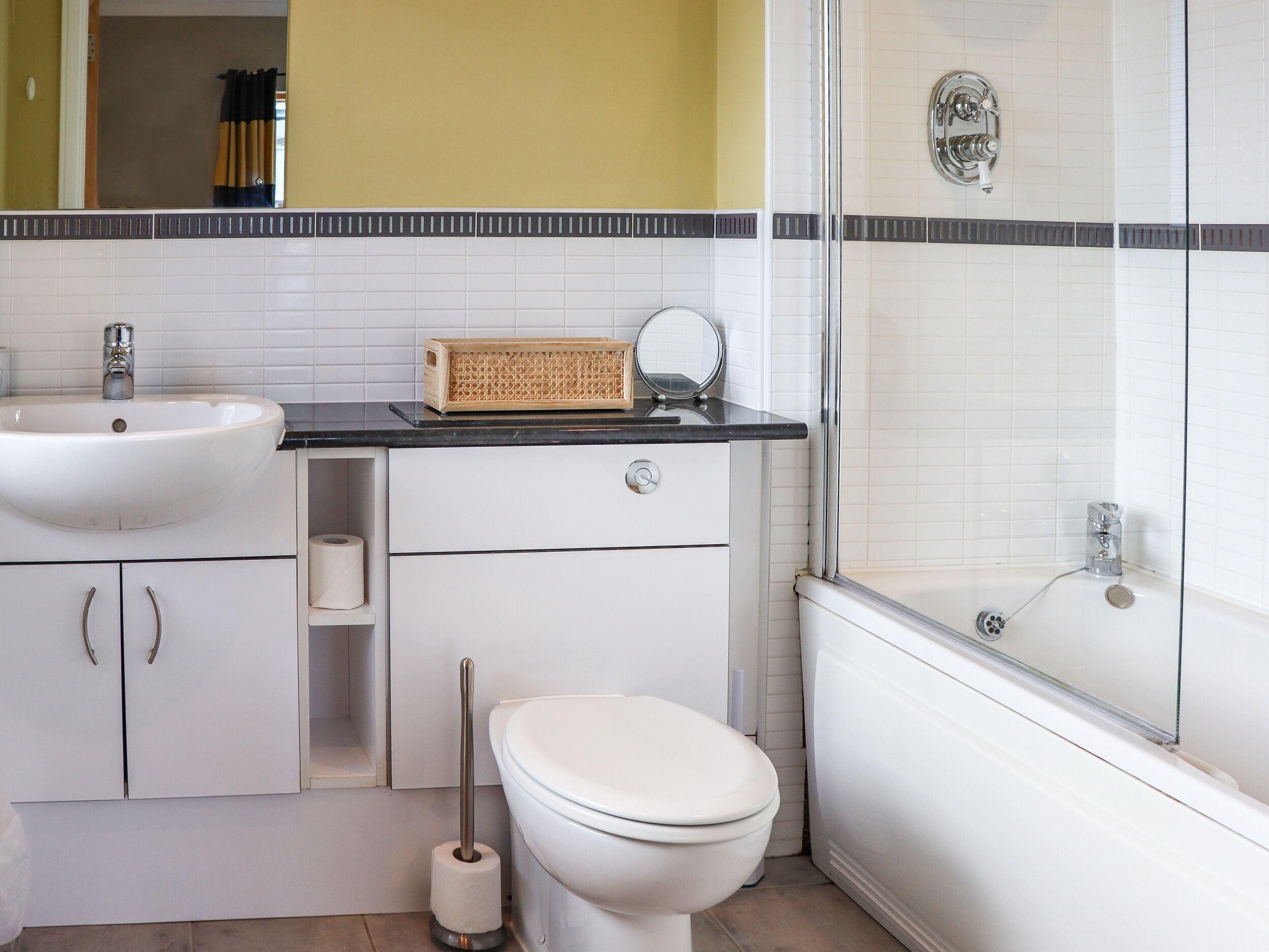 A bathroom with a white sink, toilet, and shower. There is a mirror on the countertop and a toilet paper roll in the cabinet.