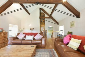 Ein geräumiges Wohnzimmer mit hoher Decke und freiliegenden Holzbalken, möbliert mit Ledersofas und einem Holztisch.