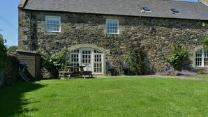 A stone building with a sloped roof, a small garden with a picnic table, and a wooden shed. - Budle Sands (Bamburgh)