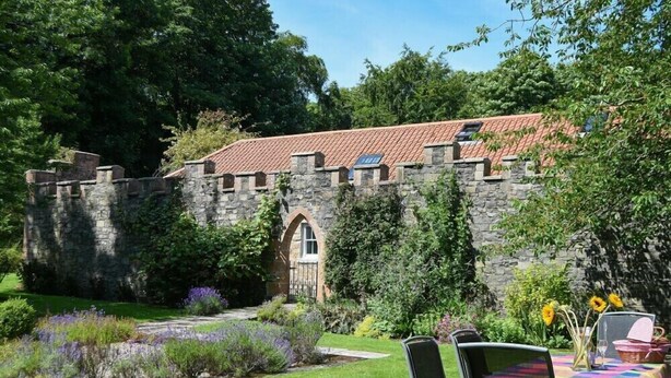 A stone building with a red-tiled roof, surrounded by lush greenery and a garden with purple flowers.