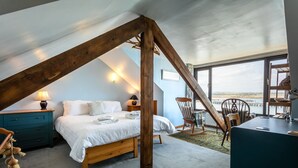 Een slaapkamer met een schuine kap, een bed, een houten schommelstoel, een bureau en uitzicht op het water door grote ramen.