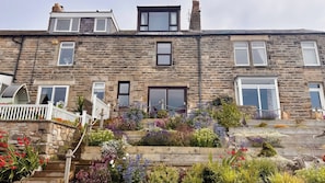 A stone-built residential building with multiple windows, a front garden with various plants, and a white fence. - Old Coastguard Cottages (Morpeth)