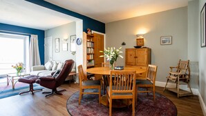 Een gezellige woonkamer met een houten eettafel, comfortabele zitplaatsen, een boekenkast en een groot raam met gordijnen.