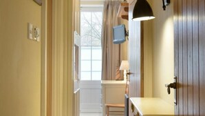 Ein schmaler Flur mit einer Holztür rechts, einem kleinen Tisch mit Lampe und einem Fenster mit Vorhängen.