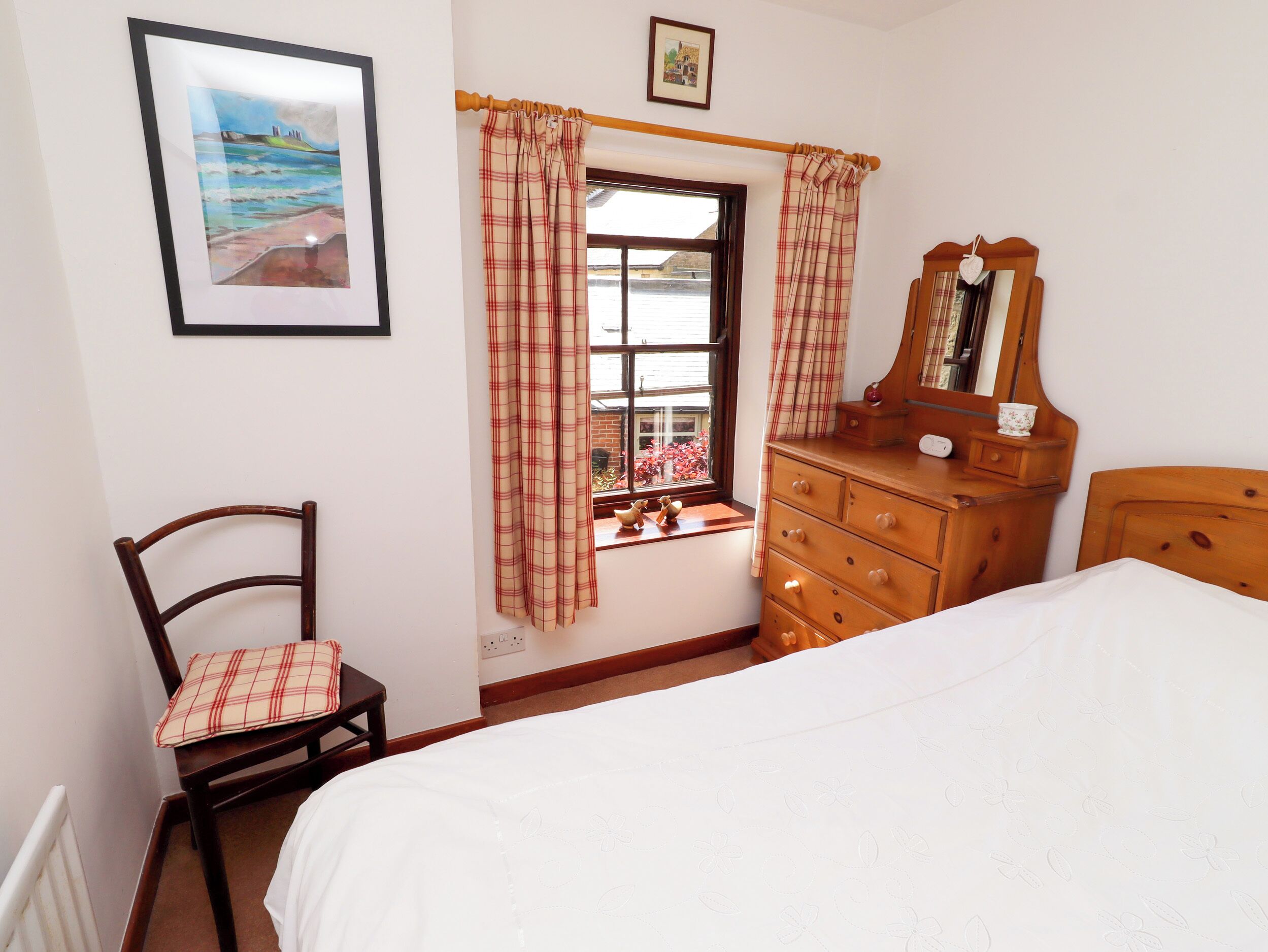 Een slaapkamer met een bed, een kaptafel, een stoel met een geruit kussen, een raam met gordijnen en een ingelijste foto aan de muur.