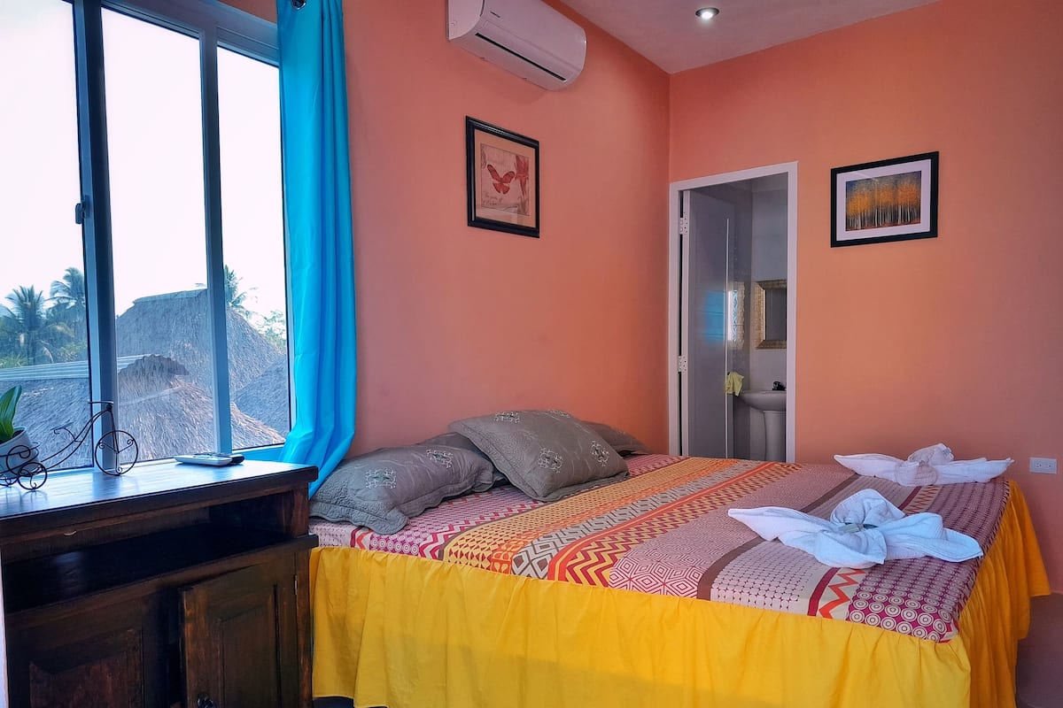 A bedroom with a bed, a window with curtains, a bedside table, and a door leading to a bathroom.