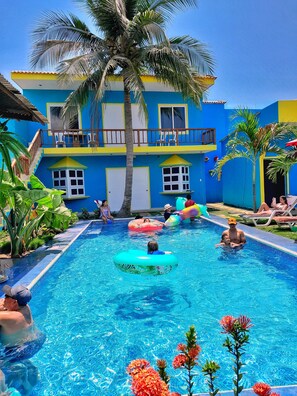Un espace piscine avec un bâtiment bleu, des palmiers et des personnes utilisant des bouées gonflables.