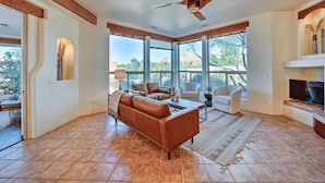 A spacious living room with a large sofa, a coffee table, and a TV. There are large windows providing a view of the outdoors. - Red Rock Mtn View & Fire Pit: Central Sedona Home! (Sedona)
