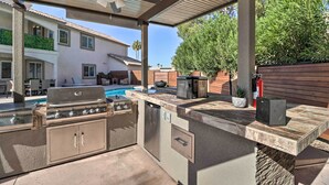 Une cuisine extérieure équipée d’un barbecue, d’un évier et de placards de rangement, donnant sur un jardin avec une piscine et des meubles de patio.