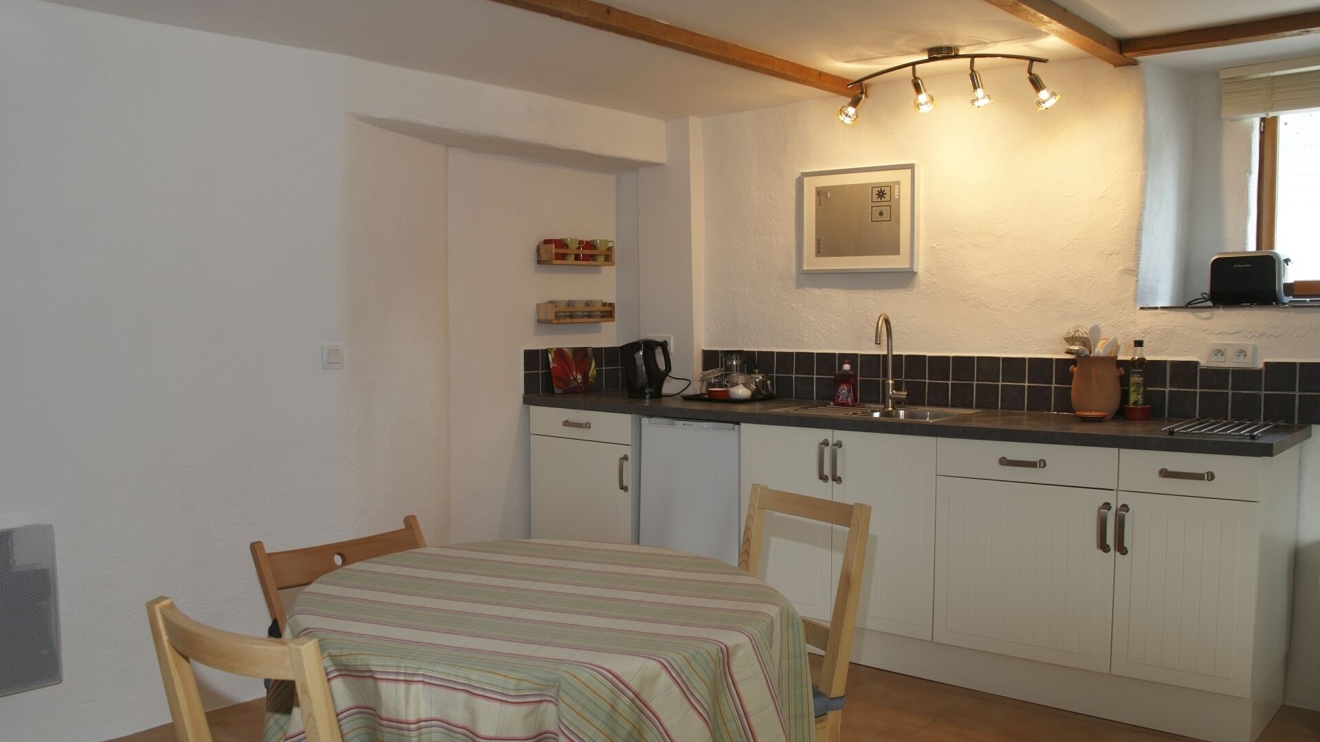 Een kleine keuken met witte kasten, een zwart aanrecht en een eettafel met stoelen.