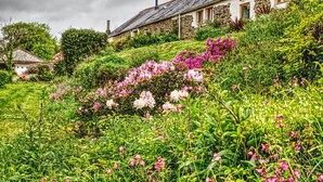 A stone house with a sloped roof, surrounded by lush greenery and flowering plants.