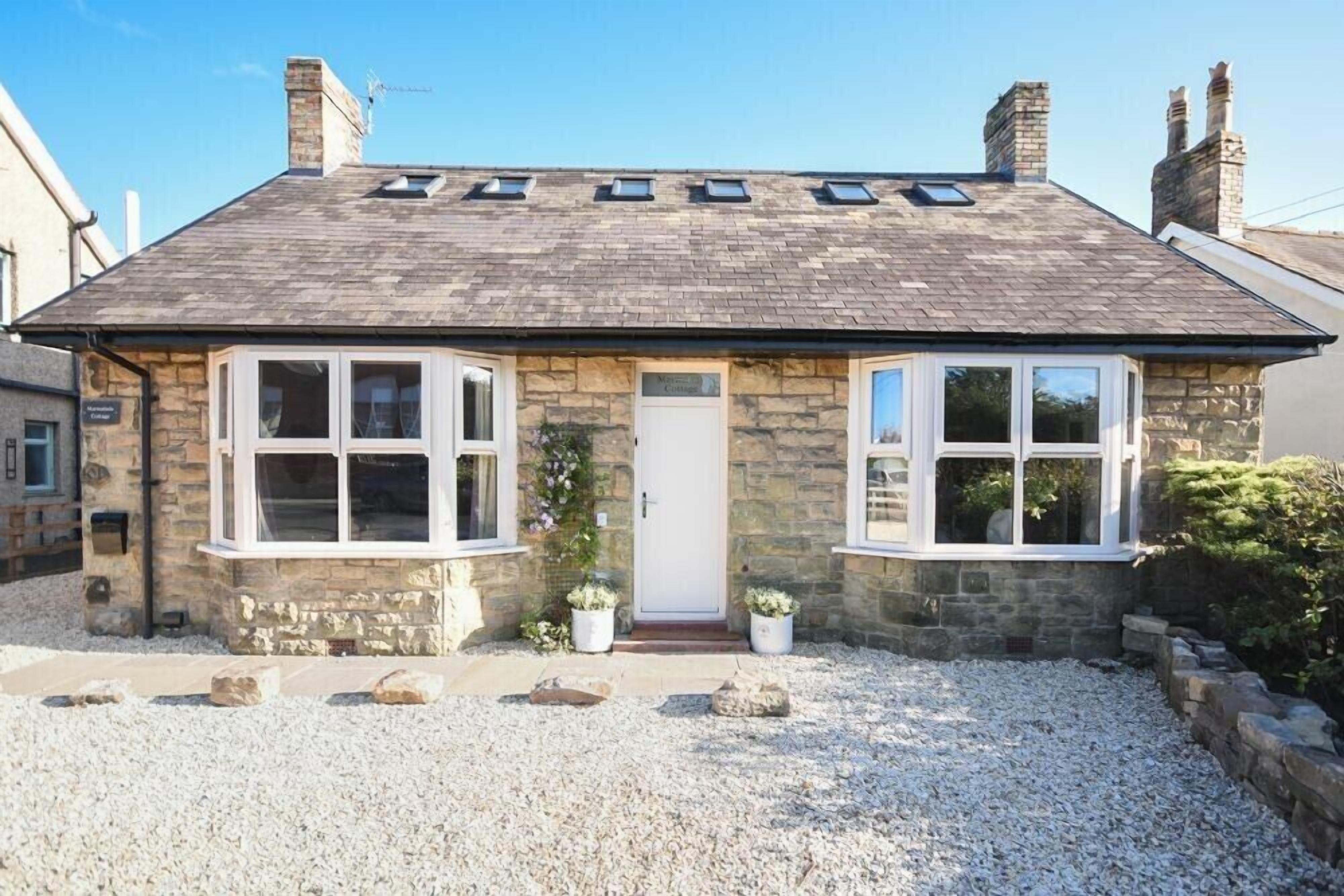 A stone cottage with a sloped roof, two bay windows, a front door, and a gravel driveway.