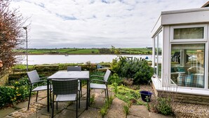 A patio with a table and chairs, a stone pathway, and a view of a lake with boats. - The Beach House, Alnmouth (Alnwick)