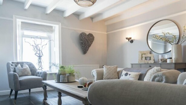 A living room with a grey sofa, a matching armchair, a wooden coffee table, and a heart-shaped wall decoration. - Creel Cottage, Craster (Alnwick)