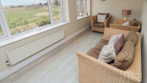 A spacious living room with a large window offering a view of a grassy field. There are two wicker sofas with cushions and a small table with a lamp. - Beach View (Seahouses)