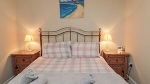 A neatly made bed with a checkered bedspread, two pillows, and rolled towels on a headboard. There are two bedside tables with lamps and a clock. A painting of a beach scene hangs above the bed. - Beach View (Seahouses)