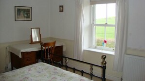 Een slaapkamer met een bed, een houten stoel, een dressoir, een spiegel en een raam met uitzicht op een groen veld.