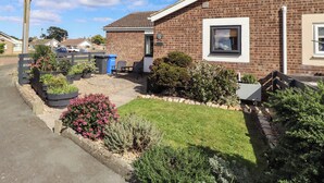 A residential house with a brick exterior, a front garden with various plants, a visible satellite dish, and a clear sky.