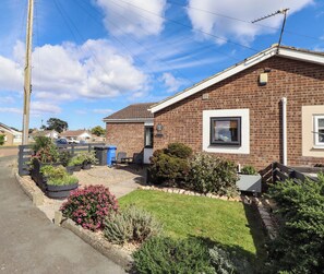 A residential house with a brick exterior, a front garden with various plants, a visible satellite dish, and a clear sky.