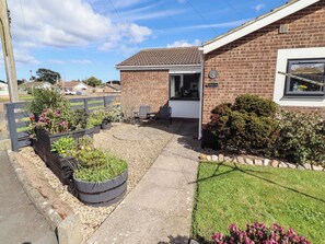 A residential property with a brick exterior, a small garden with raised flower beds, a paved driveway, and a door leading to the entrance.