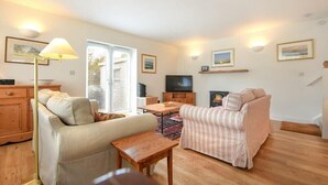 A cozy living room with a sofa, a striped armchair, a wooden coffee table, and a TV. There are framed pictures on the walls and a fireplace. - Stone Cottage (Bamburgh)