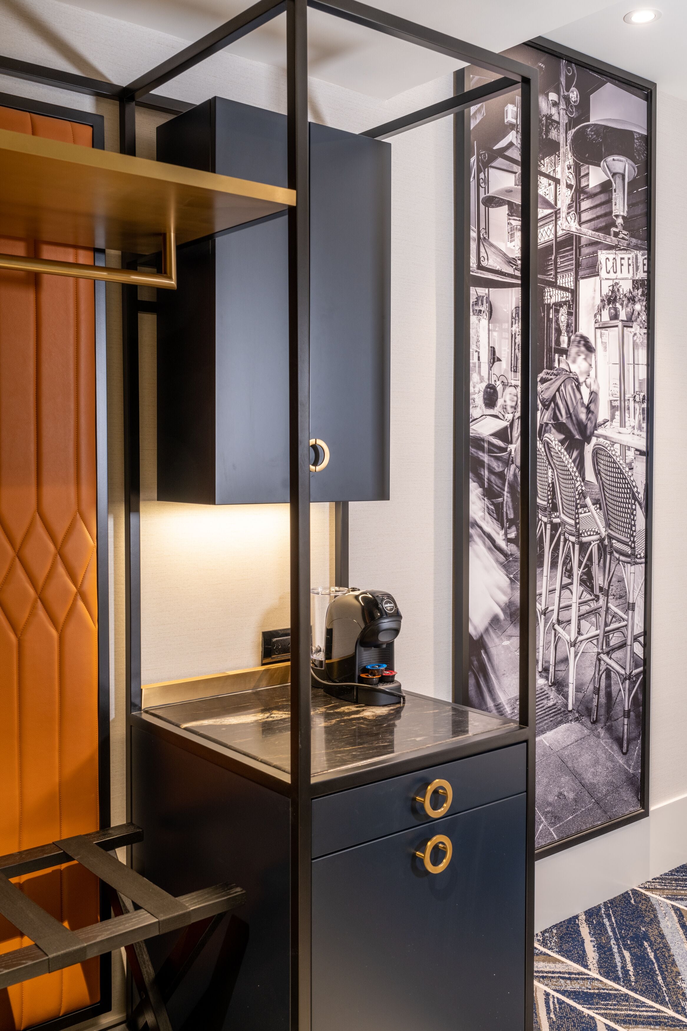 A modern, compact room with a black and gold color scheme, featuring a desk with a coffee machine, a framed black and white photograph on the wall, and a patterned rug.