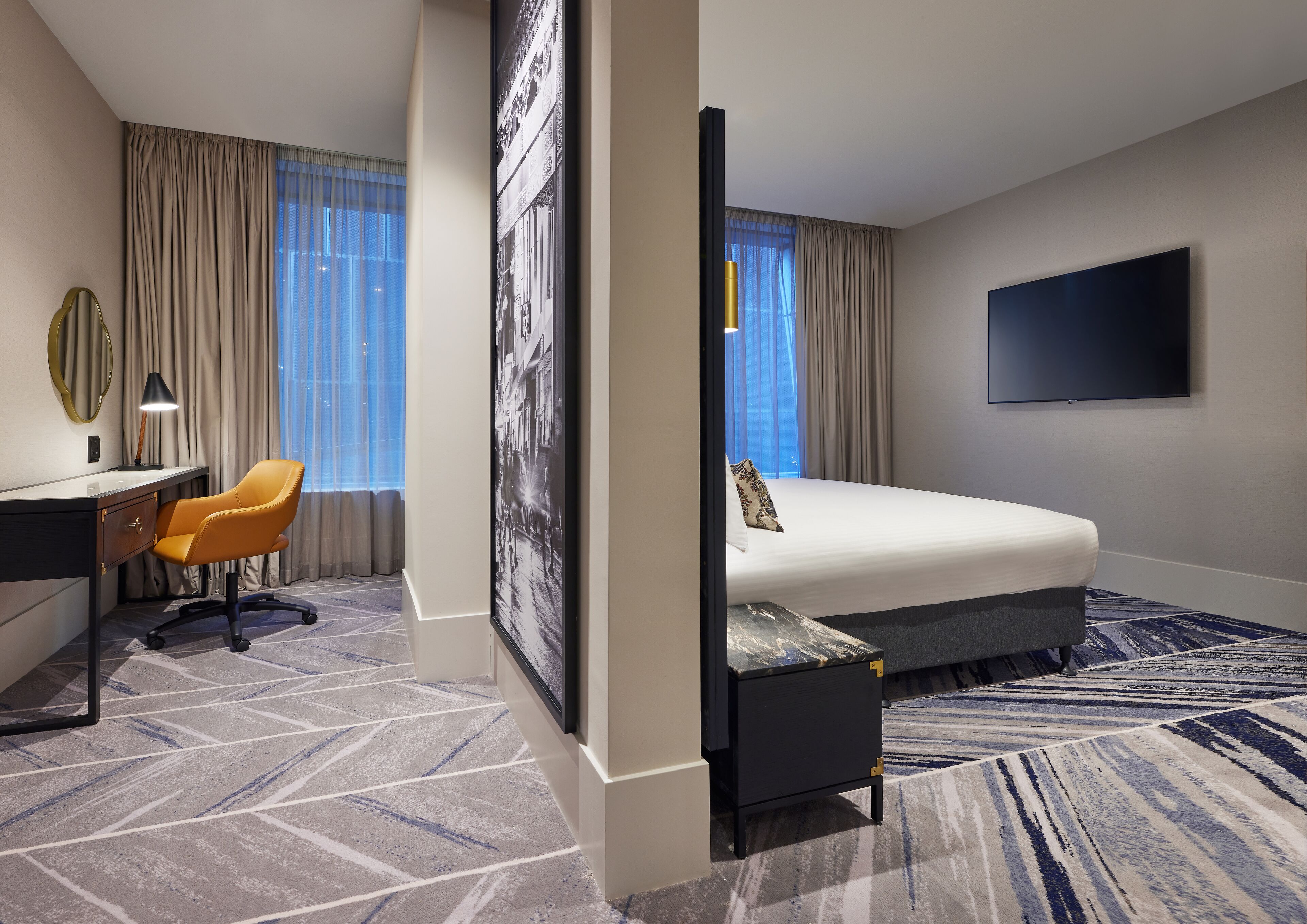 A hotel room with a bed, desk, chair, and a large TV on the wall.