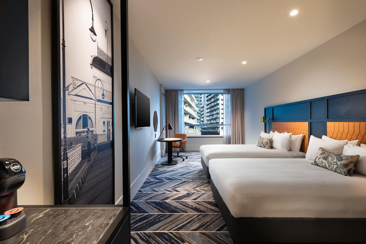 A hotel room with two beds, a desk, a chair, a TV, and a view of high-rise buildings.