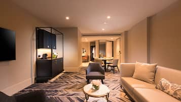 A modern hotel room with a sofa, armchair, and a small round table with a vase. There is a kitchen area with cabinets and a dining table.