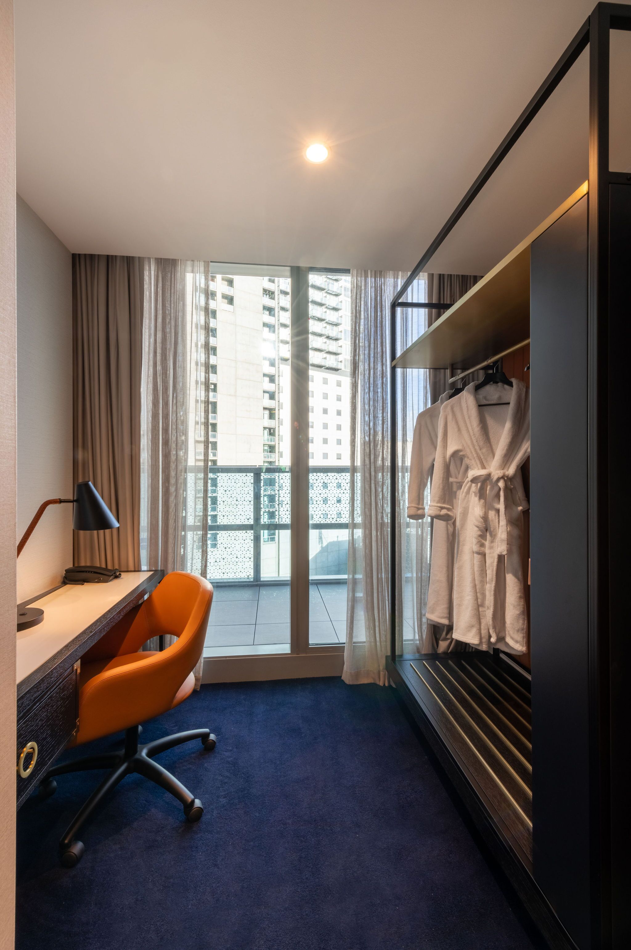 Quarto de hotel com escrivaninha, uma cadeira laranja, guarda-roupa e vista para edifícios altos pela janela.
