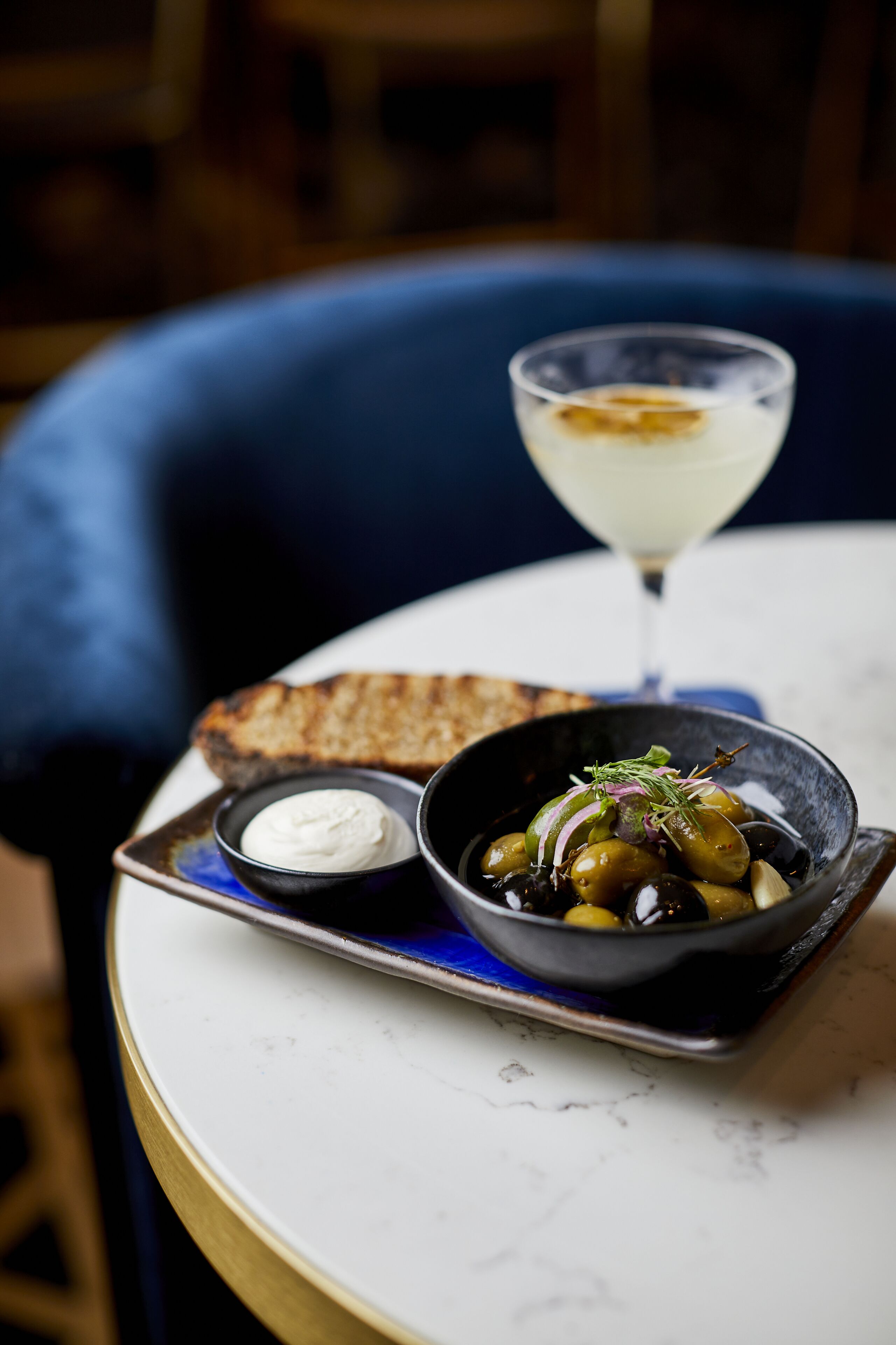 A table with a bowl of olives, a small dish of cream, and a glass of white wine.