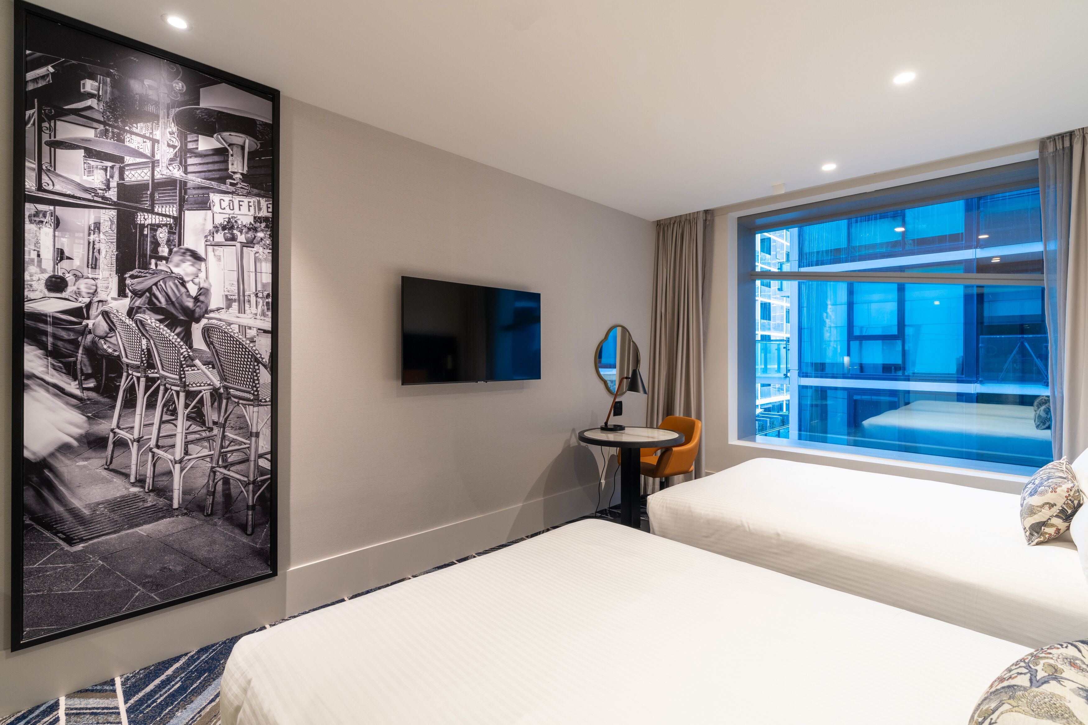 A hotel room with a bed, a TV, a small table with a chair, a large window, and a wall-mounted mirror.