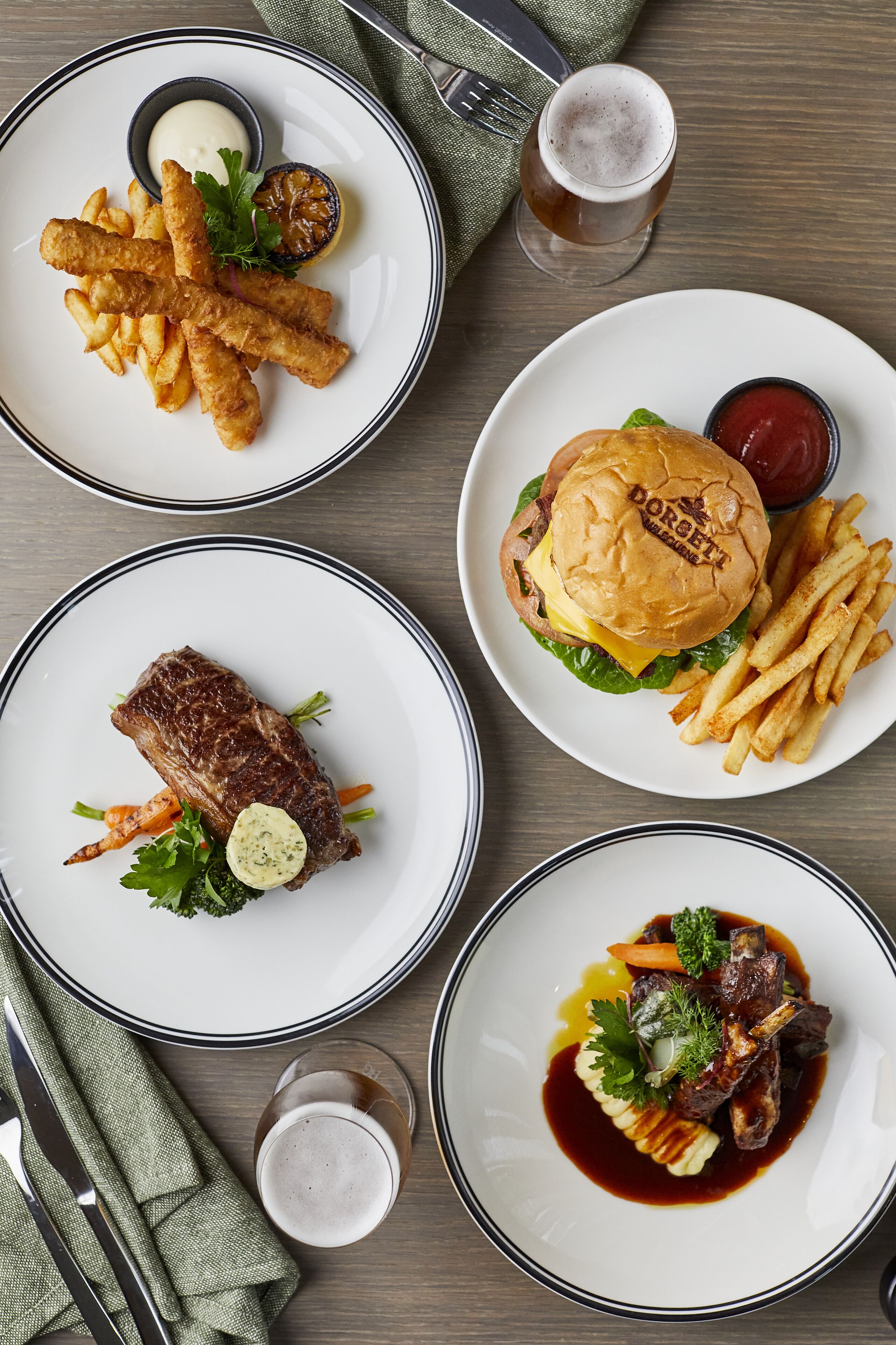 Una mesa con cuatro platos de comida, incluyendo una hamburguesa, papas fritas y un bistec, junto con dos vasos de cerveza.