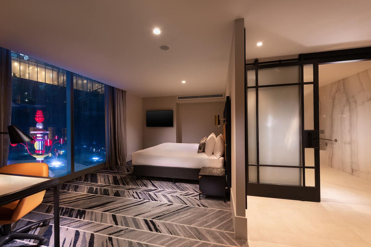 A modern hotel room with a large bed, a desk with a chair, a TV mounted on the wall, and a view of the city at night through large windows.